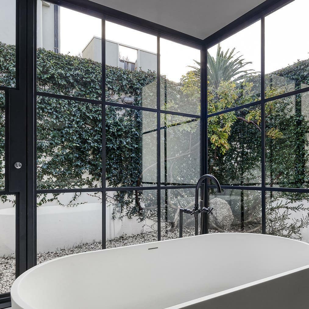 corner windows with bathtub, looking out onto wall with vines growing over it. the windows have black panel framing and a door to the outdoor area